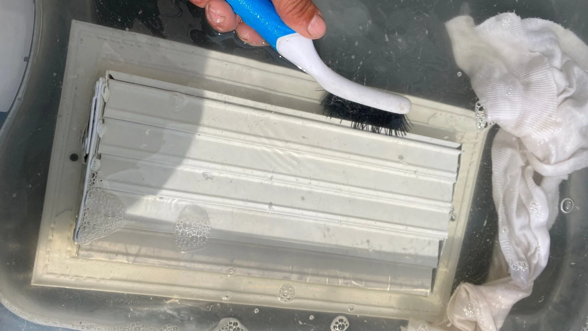 Hand scrubbing a removed white metal air register in a tub with water and suds.